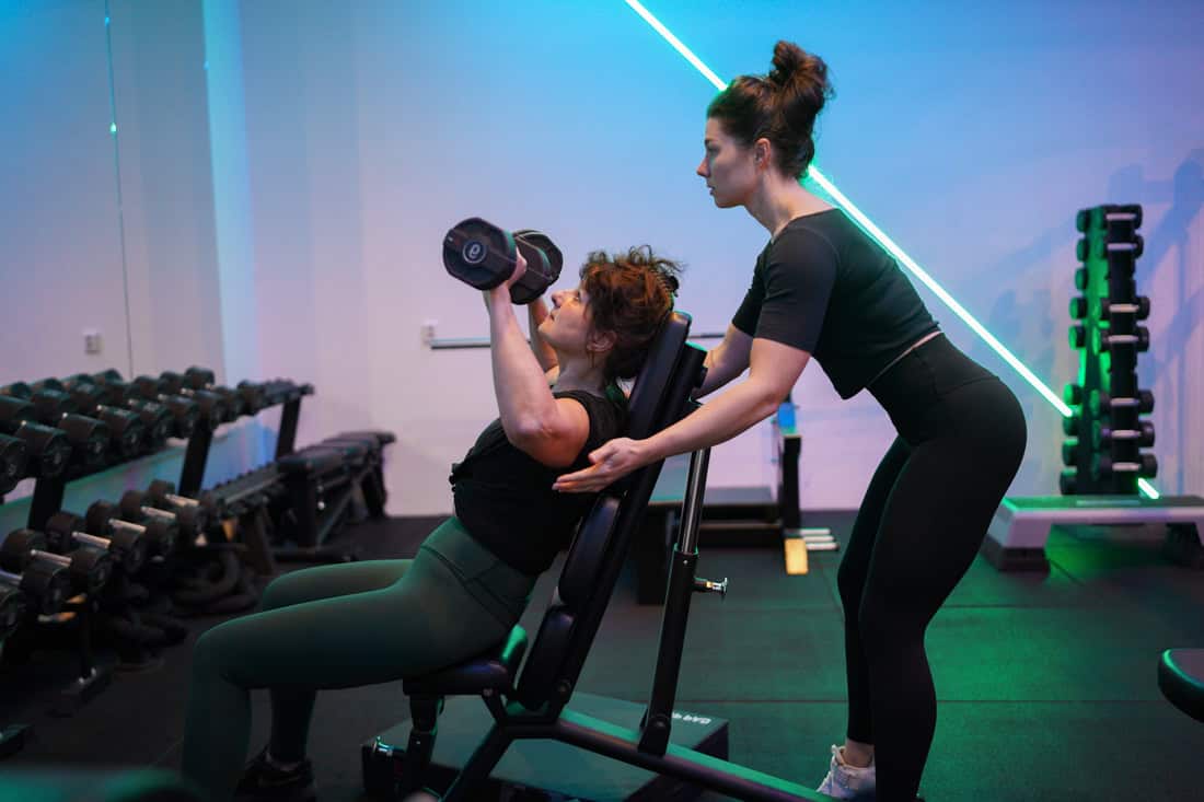 Anna begeleidt een vrouw bij een shoulder press met dumbbells.